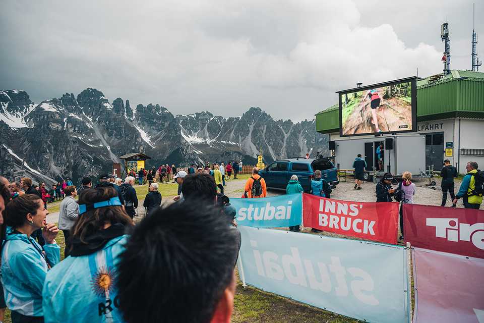 Trail Run WM Innsbruck-Stubai: Zuschauer verfolgen Rennen auf Leinwand vor Bergkulisse.