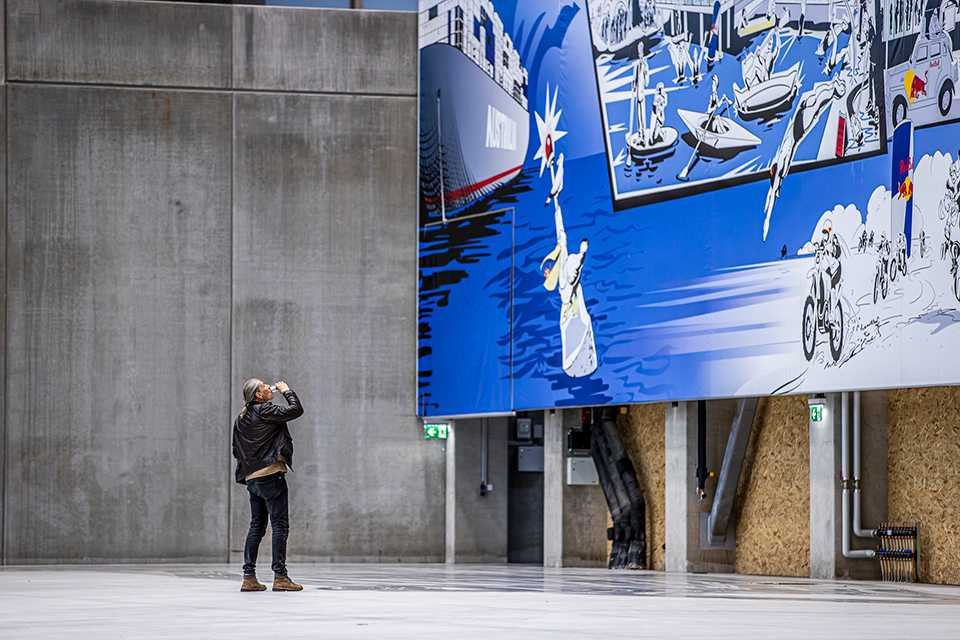 Mann betrachtet Red Bull Energy Station Kunst in Oberwang. Wandbild mit Schiff, Sportarten und Red Bull Logo.